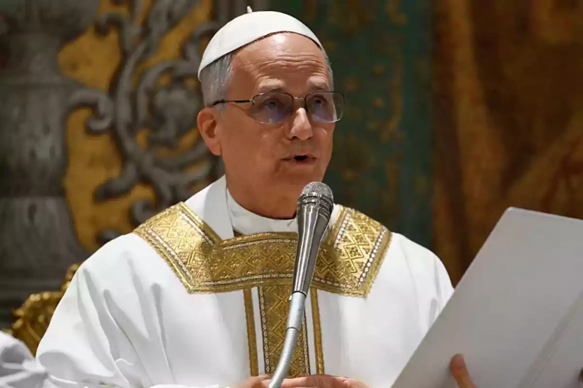 A Catholic priest in white and gold liturgical vestments speaks into a microphone while holding a document. A Catholic priest in white and gold liturgical vestments speaks into a microphone while holding a document.