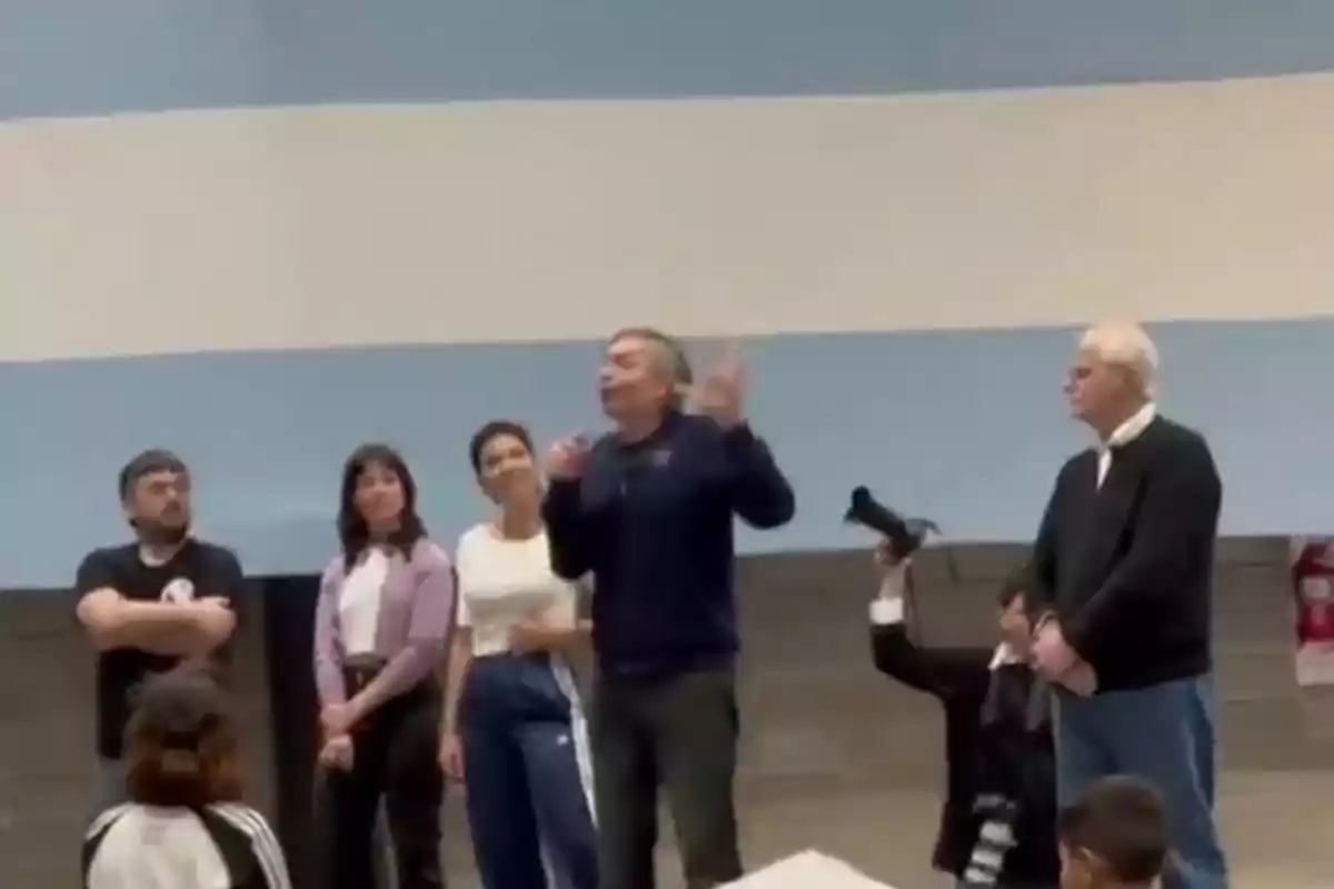 Cinco adultos están de pie en un escenario frente a un grupo de personas, uno de ellos habla con un micrófono mientras los demás observan y un niño sostiene una cámara. Cinco adultos están de pie en un escenario frente a un grupo de personas, uno de ellos habla con un micrófono mientras los demás observan y un niño sostiene una cámara.