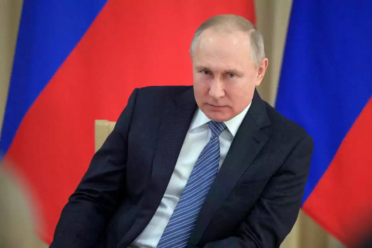 Man in a dark suit and blue tie sitting in front of flags with red and blue colors Man in a dark suit and blue tie sitting in front of flags with red and blue colors