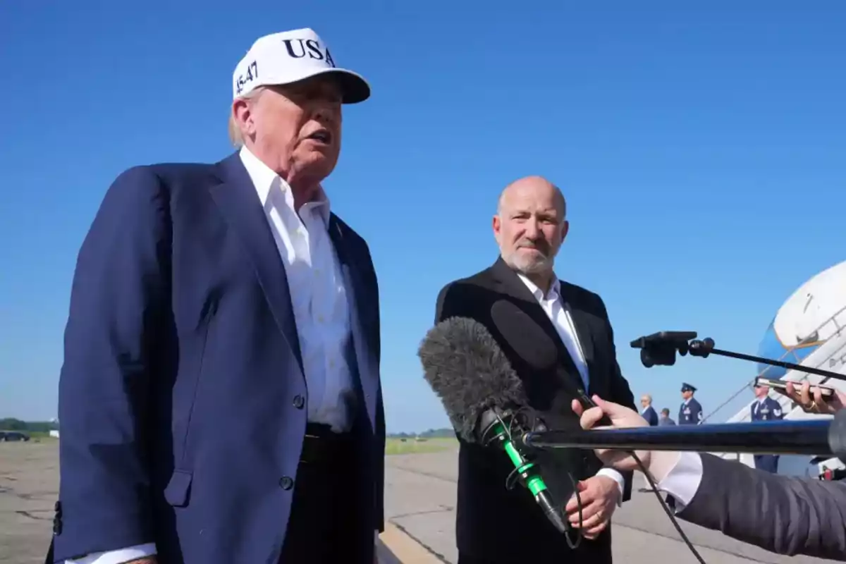 Two men in suits are outdoors on a runway while answering questions in front of several microphones with a clear sky in the background.
