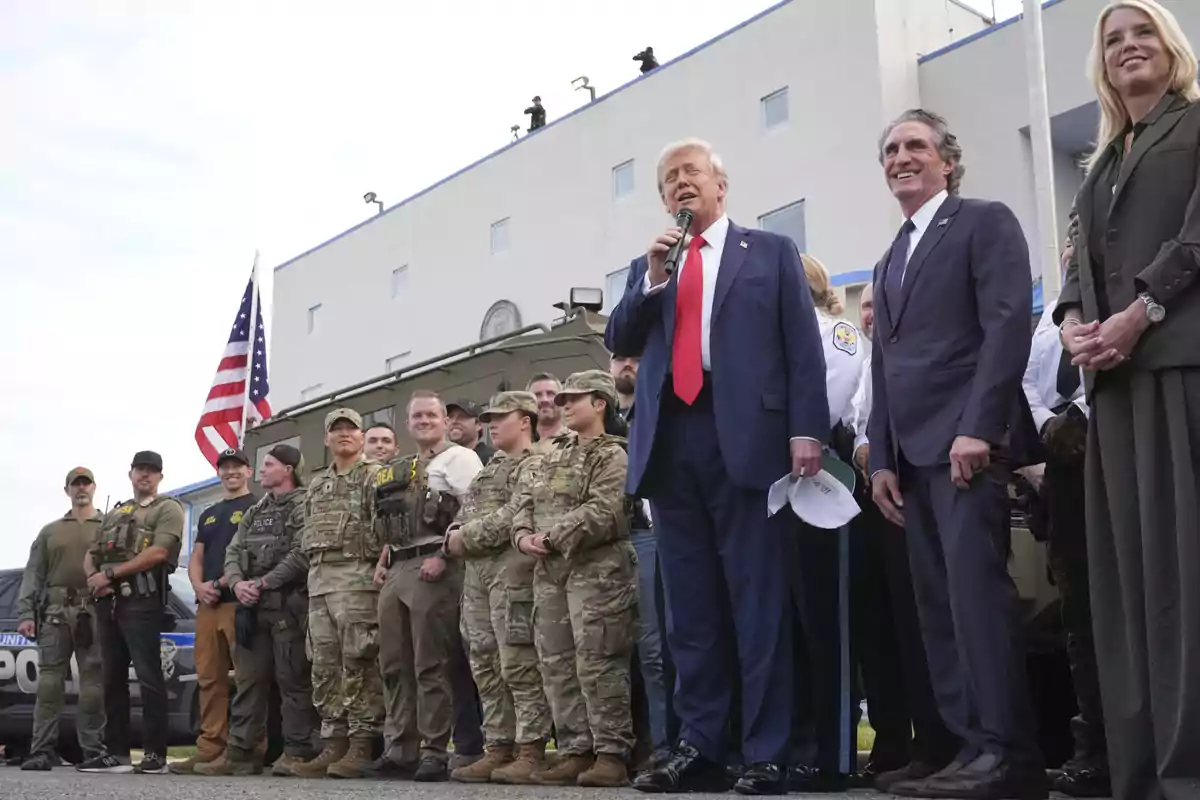 Un grupo de militares y policías posan junto a un hombre de traje azul con corbata roja que sostiene un micrófono, acompañados de otras personas frente a un edificio y una bandera de Estados Unidos