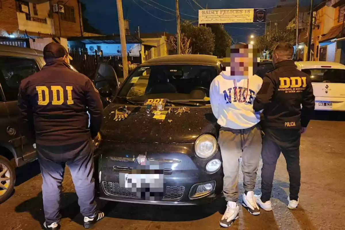 Two police officers wearing jackets that say DDI detain a person in front of a black car on a street at night, with tools and gloves on the hood of the vehicle.