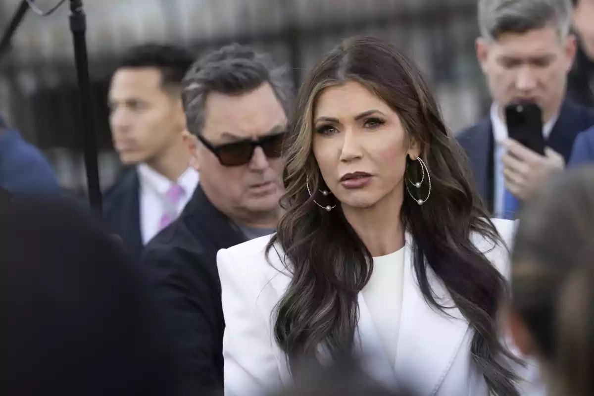 Mujer de cabello largo y oscuro con chaqueta blanca y aretes grandes rodeada de varias personas en un evento al aire libre Mujer de cabello largo y oscuro con chaqueta blanca y aretes grandes rodeada de varias personas en un evento al aire libre