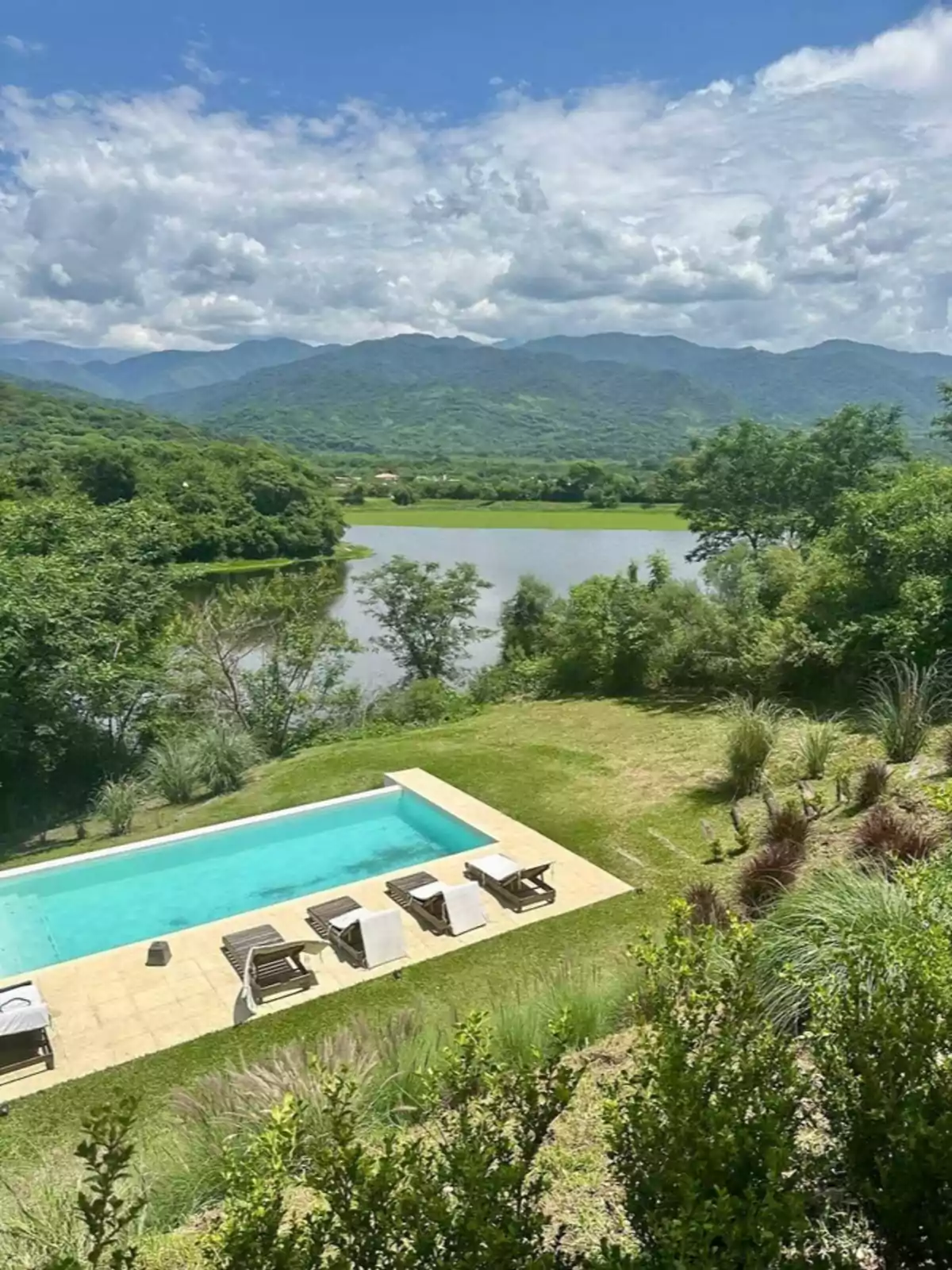 Piscina al aire libre rodeada de césped con varias reposeras y toallas, vista a un lago y montañas cubiertas de vegetación bajo un cielo parcialmente nublado Piscina al aire libre rodeada de césped con varias reposeras y toallas, vista a un lago y montañas cubiertas de vegetación bajo un cielo parcialmente nublado