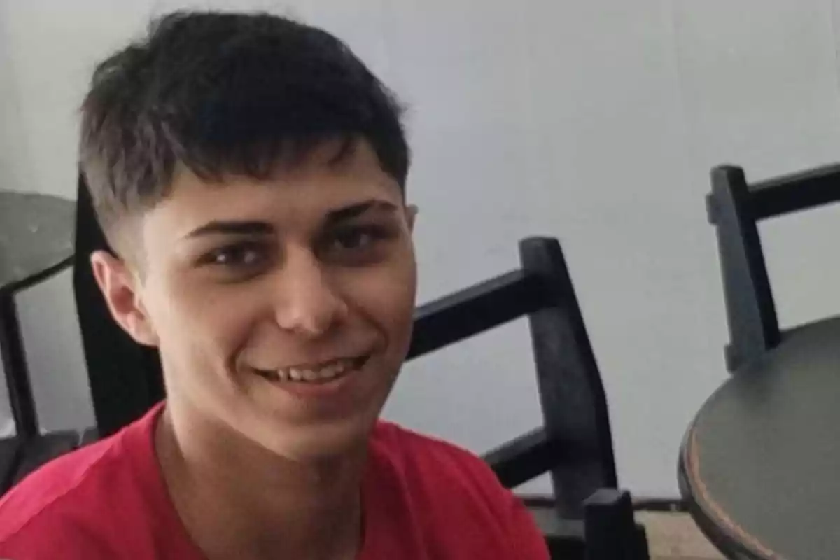 Young man with short dark hair smiles while sitting next to a table in an indoor setting. Young man with short dark hair smiles while sitting next to a table in an indoor setting.