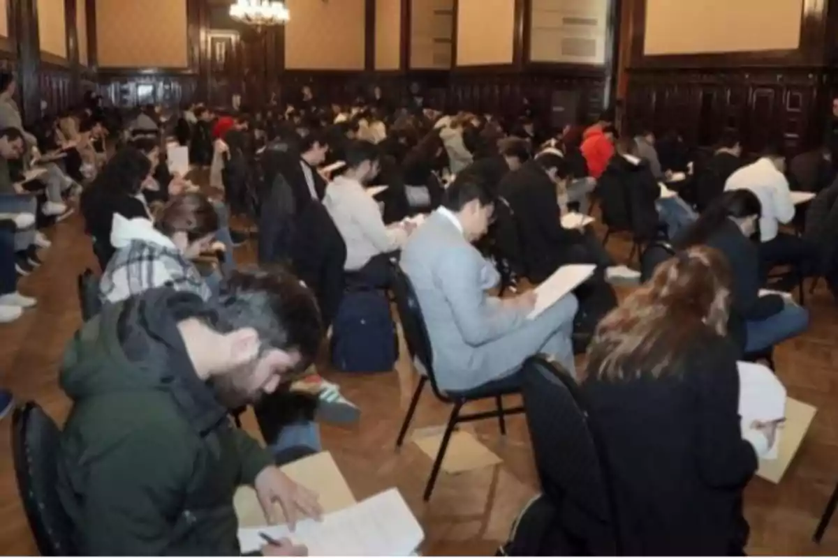 Personas sentadas en un salón grande realizando un examen escrito, concentradas en sus hojas y bolígrafos Personas sentadas en un salón grande realizando un examen escrito, concentradas en sus hojas y bolígrafos