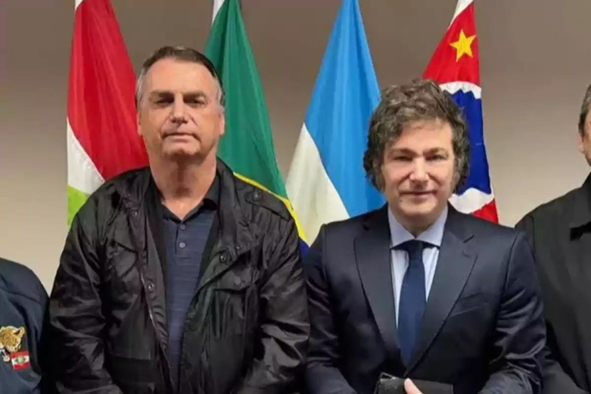 Two men pose together in front of several flags; one is wearing a black jacket and the other is dressed in a dark blue suit with a tie. Two men pose together in front of several flags; one is wearing a black jacket and the other is dressed in a dark blue suit with a tie.