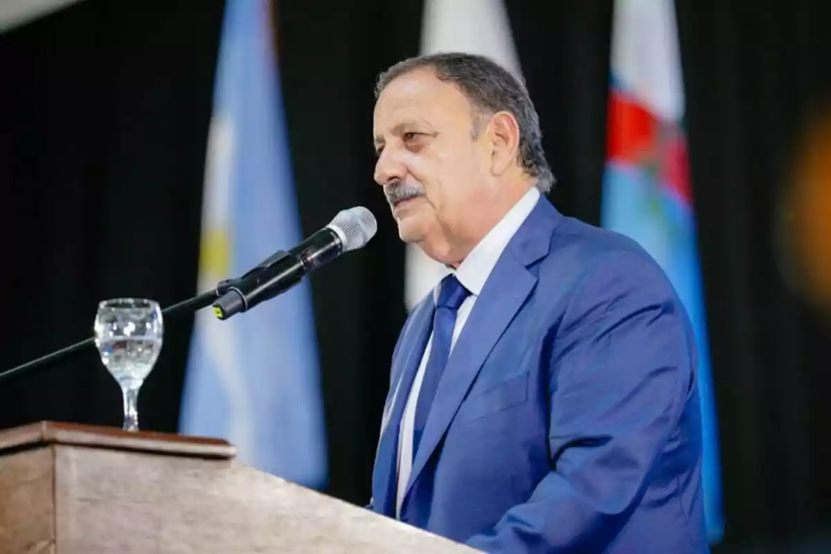 Hombre de traje azul hablando en un podio con micrófono y copa de agua, banderas desenfocadas al fondo