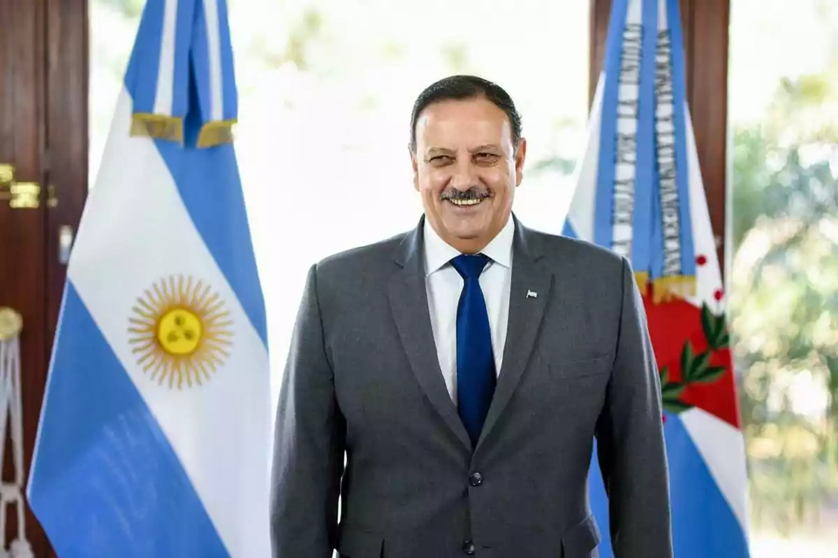 Hombre de traje gris y corbata azul sonriendo frente a dos banderas argentinas Hombre de traje gris y corbata azul sonriendo frente a dos banderas argentinas