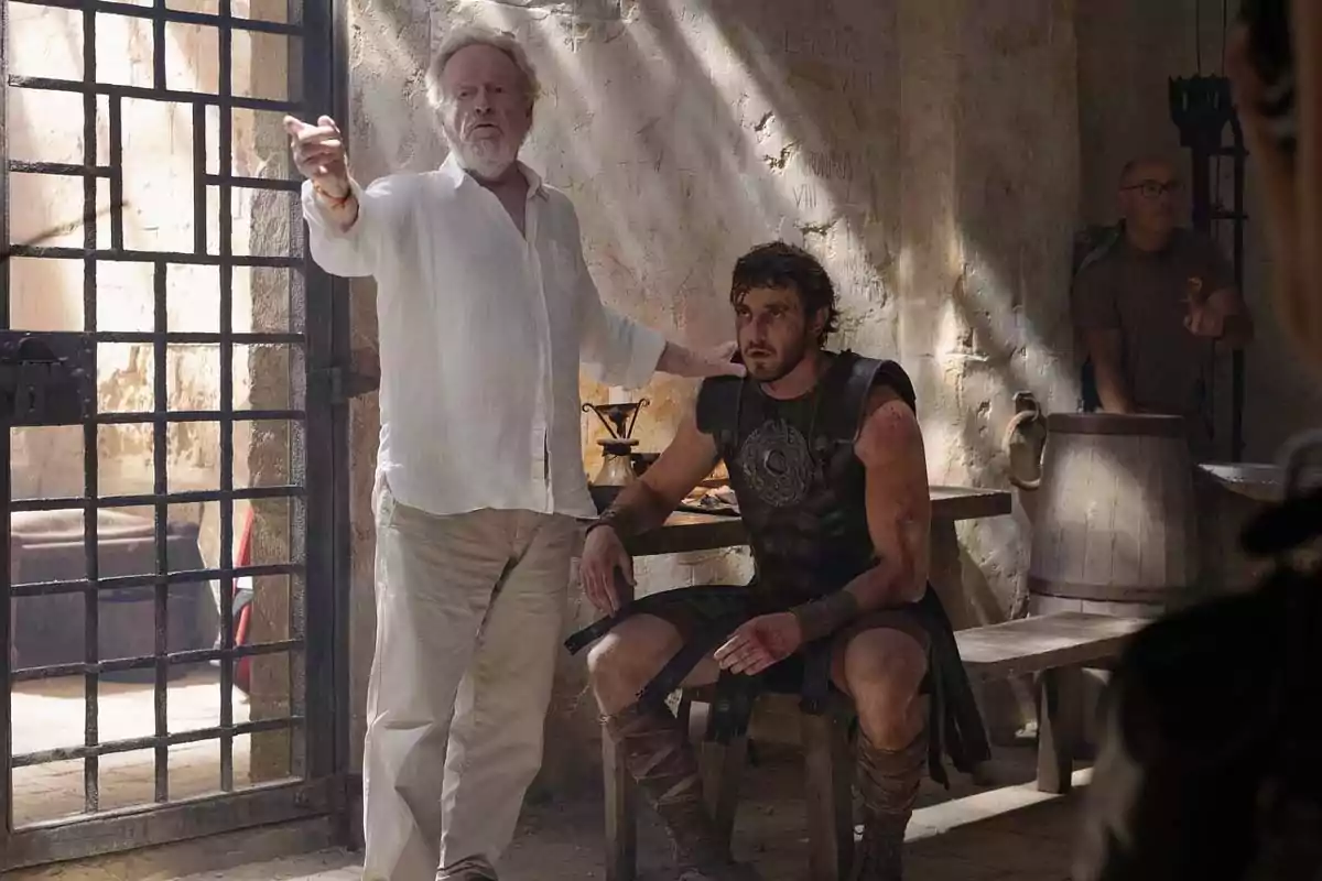 A white-haired, bearded man wearing light-colored clothes stands next to an actor dressed as a Roman warrior, who is sitting on a bench inside an old-looking room with worn walls and an iron-barred door. A white-haired, bearded man wearing light-colored clothes stands next to an actor dressed as a Roman warrior, who is sitting on a bench inside an old-looking room with worn walls and an iron-barred door.