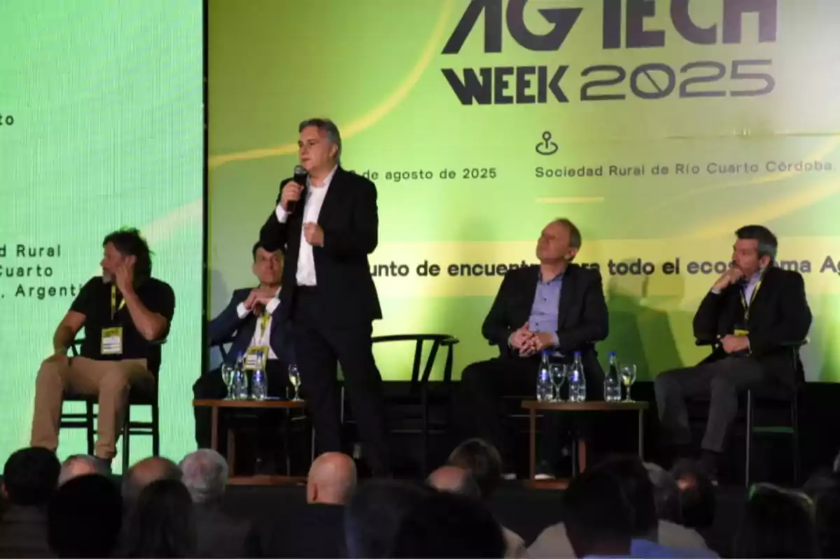 Five men participate in a panel discussion at an event called AG TECH WEEK 2025, with a green and yellow background and an audience watching. Five men participate in a panel discussion at an event called AG TECH WEEK 2025, with a green and yellow background and an audience watching.