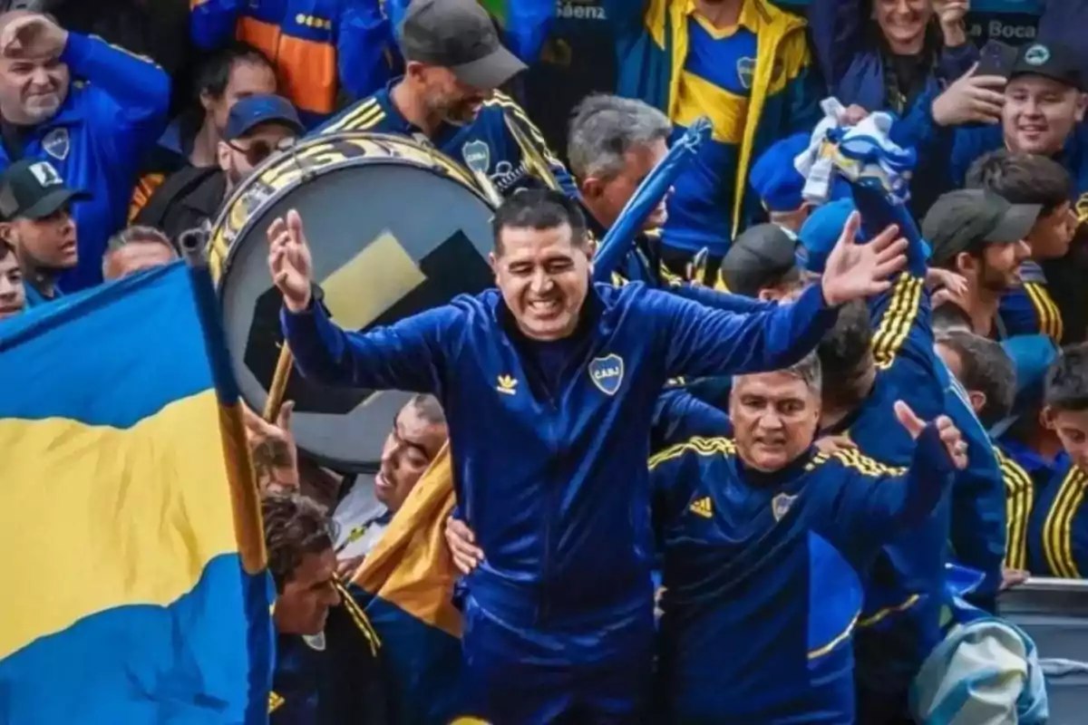 Smiling man with open arms surrounded by Boca Juniors fans with flags and drums in a festive atmosphere