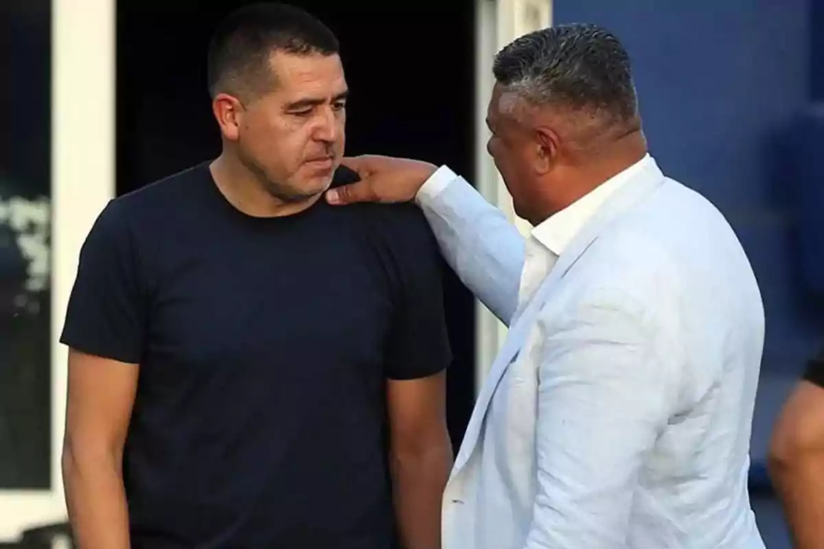 Two men talking, one wearing a black t-jersey and the other a light-colored jacket, while one rests his hand on the other's shoulder.