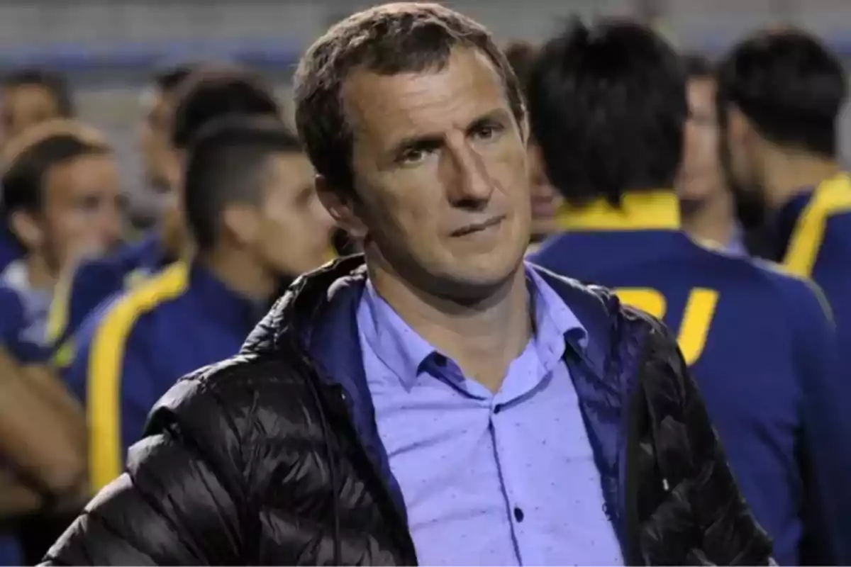 Short-haired man in a black jacket with a serious expression in a sports setting, with people in the background wearing blue and yellow uniforms.
