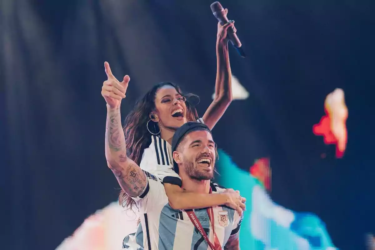 Dos personas sonrientes con camisetas de la selección argentina celebran en un escenario, una de ellas sostiene un micrófono y abraza a la otra desde la espalda