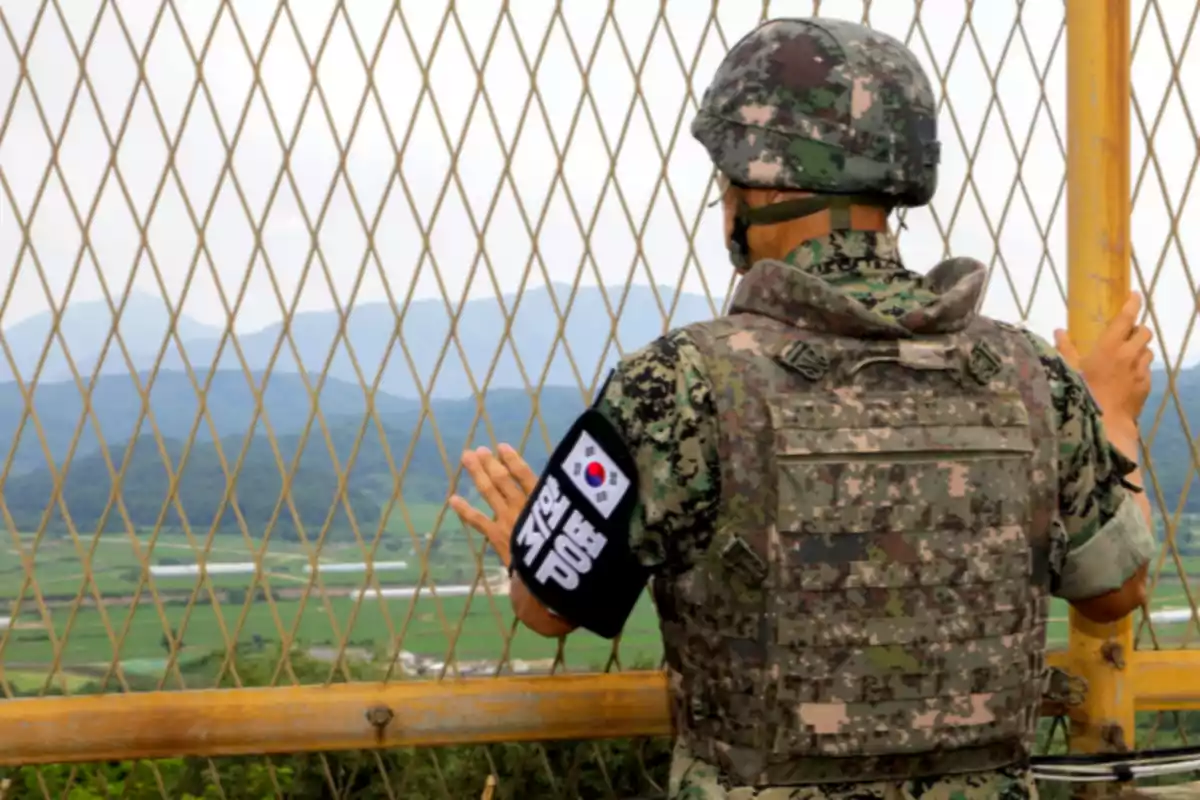 South Korean soldier in camouflage uniform looking through a metal fence at a landscape of mountains and green fields