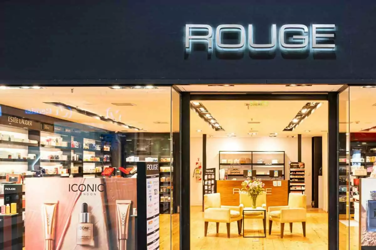 Facade of an elegant perfumery with the illuminated ROUGE sign and a well-decorated interior featuring beauty products and two armchairs next to a table with flowers. Facade of an elegant perfumery with the illuminated ROUGE sign and a well-decorated interior featuring beauty products and two armchairs next to a table with flowers.
