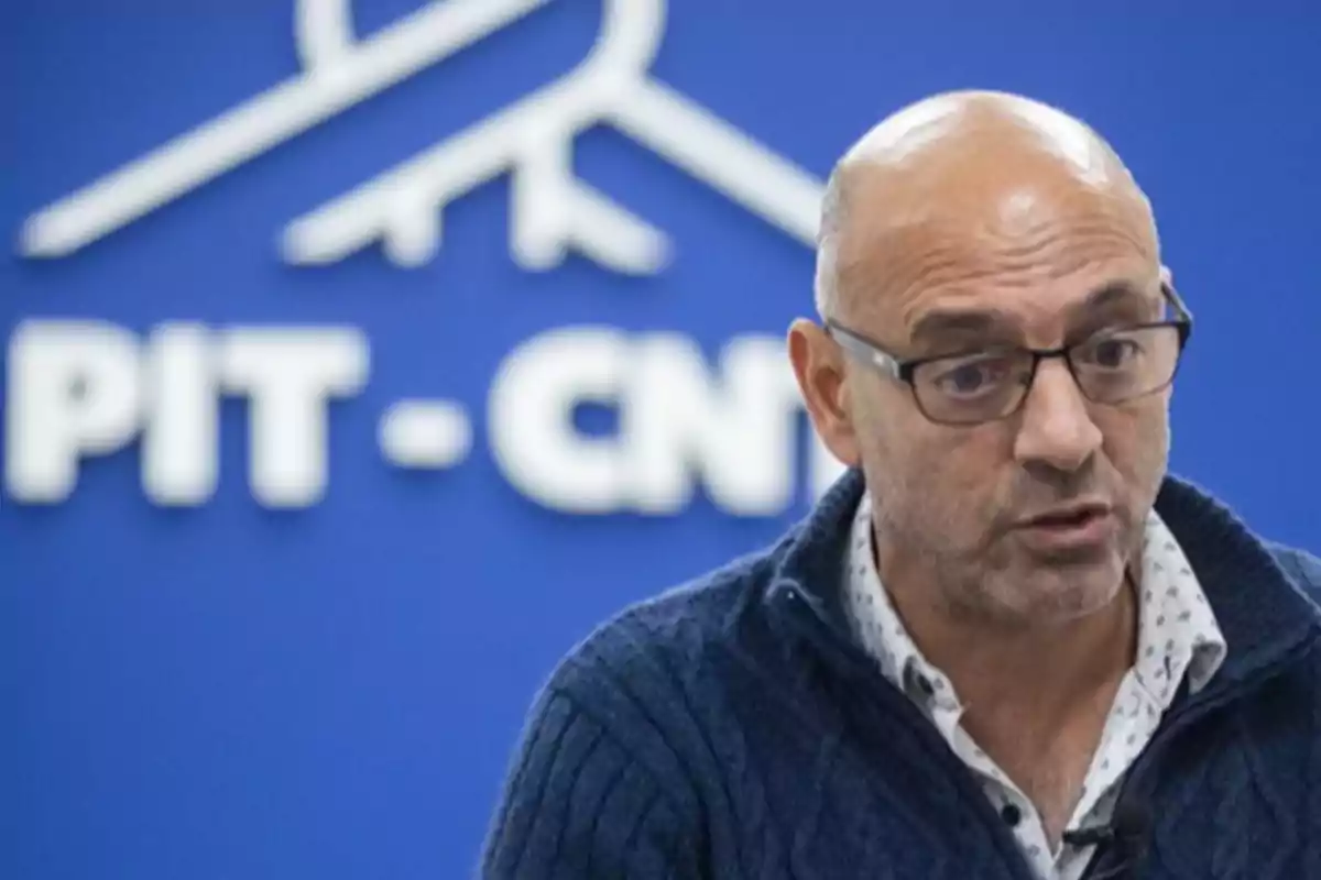 Hombre calvo con gafas hablando frente a un fondo azul con el logo y las letras PIT-CNT desenfocadas.