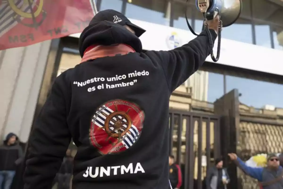 Persona de espaldas con sudadera negra que sostiene un megáfono frente a un edificio durante una manifestación la sudadera tiene la frase Nuestro único miedo es el hambre y el logo de SUNTMA Persona de espaldas con sudadera negra que sostiene un megáfono frente a un edificio durante una manifestación la sudadera tiene la frase Nuestro único miedo es el hambre y el logo de SUNTMA
