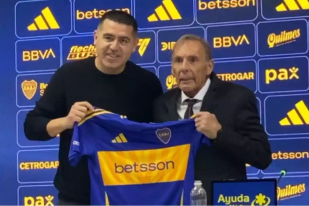 Dos hombres posan juntos sosteniendo una camiseta de Boca Juniors frente a un fondo con logotipos de patrocinadores del club. Dos hombres posan juntos sosteniendo una camiseta de Boca Juniors frente a un fondo con logotipos de patrocinadores del club.
