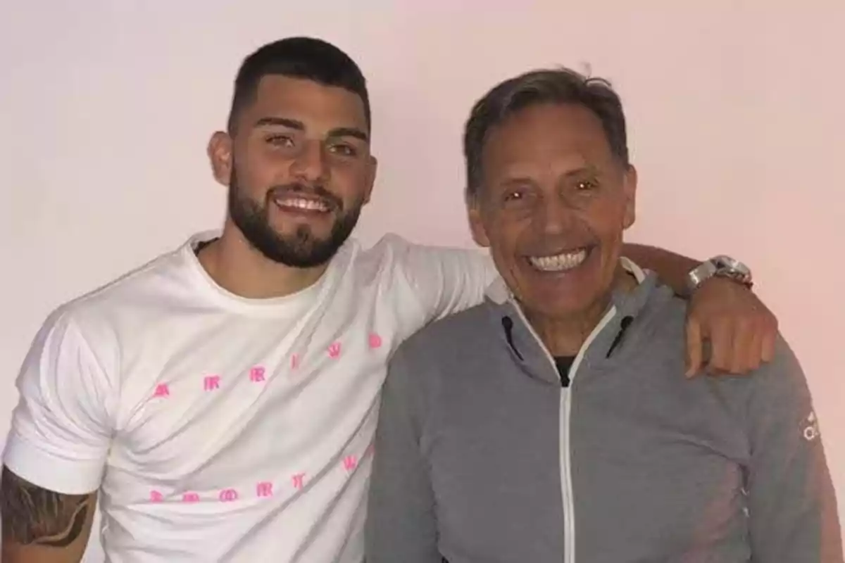 Dos hombres sonríen abrazados posando frente a una pared clara