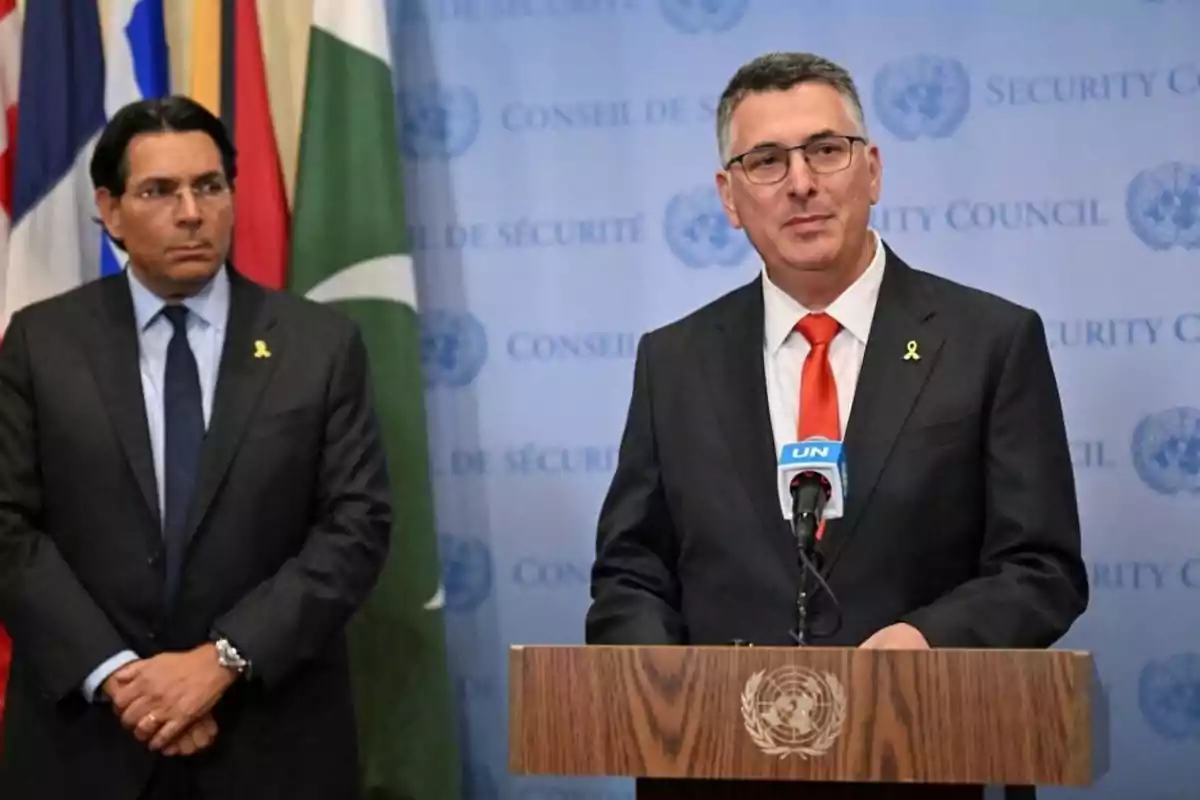 Two men in dark suits and yellow ties are at a press conference in front of a UN podium with flags of several countries in the background.