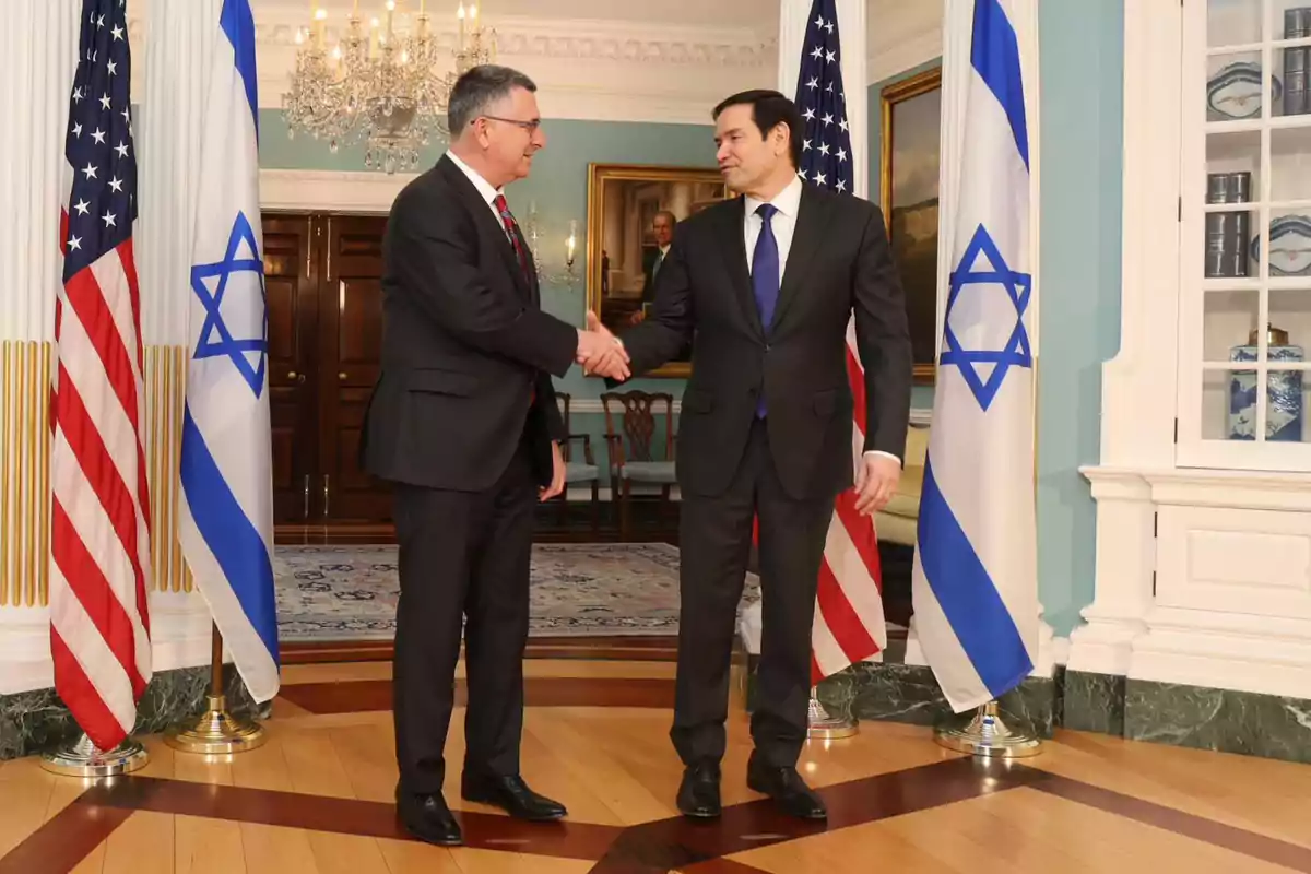 Two men in suits shake hands in an elegant room with United States and Israeli flags around them.