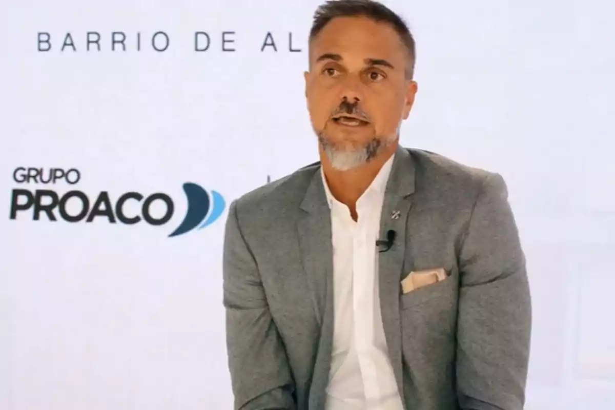 Bearded man with short hair wearing a gray jacket and white jersey sitting in front of a light background with the Grupo Proaco logo.