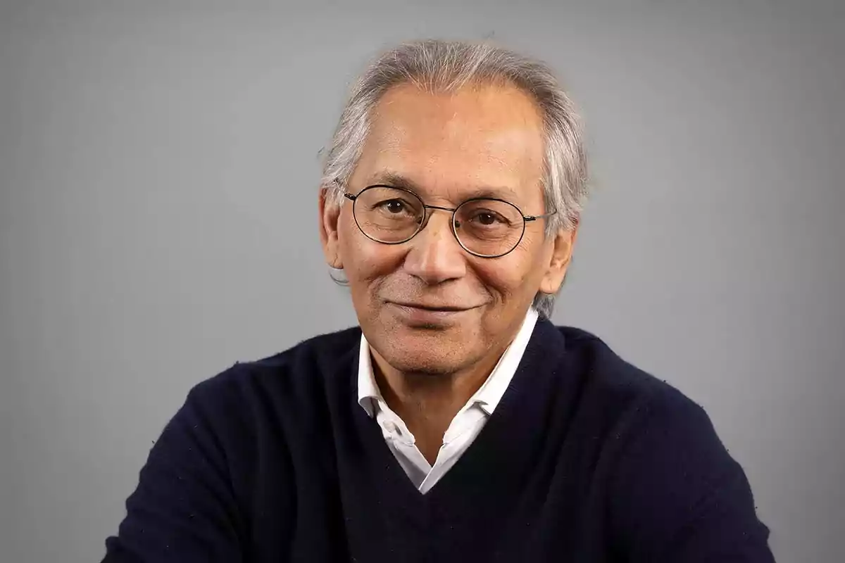Older man with gray hair and round glasses smiles slightly while wearing a navy blue sweater and a white jersey against a gray background. Older man with gray hair and round glasses smiles slightly while wearing a navy blue sweater and a white jersey against a gray background.
