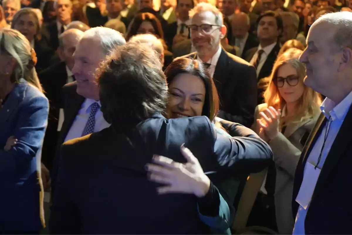 A man and a woman hug each other with smiles at an event, surrounded by people who applaud and watch with joy. A man and a woman hug each other with smiles at an event, surrounded by people who applaud and watch with joy.