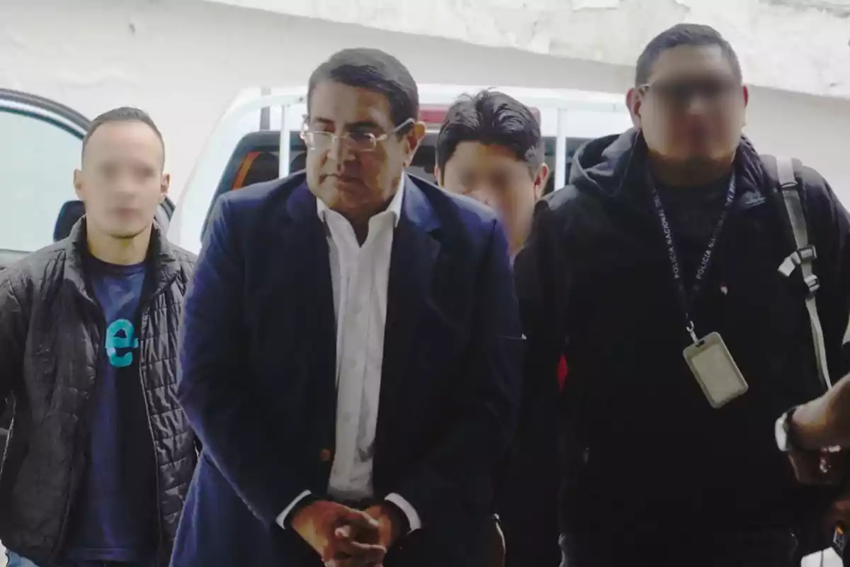 Un hombre de traje oscuro es escoltado por varias personas con el rostro difuminado mientras camina con las manos juntas frente a él.