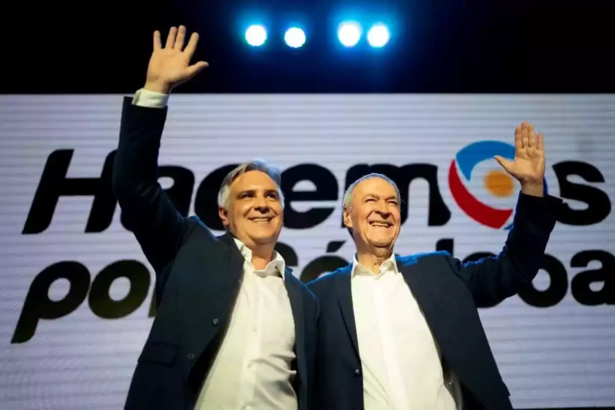 Dos hombres sonrientes con camisas blancas y sacos oscuros levantan la mano en señal de saludo frente a un cartel que dice Hacemos por Córdoba Dos hombres sonrientes con camisas blancas y sacos oscuros levantan la mano en señal de saludo frente a un cartel que dice Hacemos por Córdoba