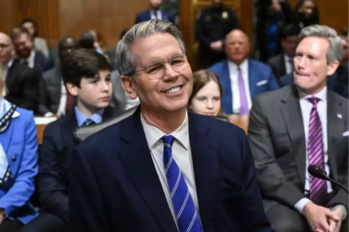 Gray-haired man with glasses smiling while sitting in a room full of formally dressed people
