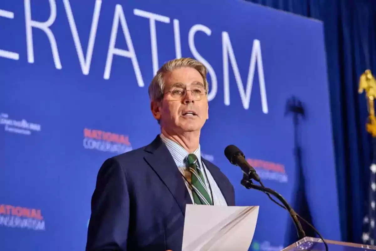 Gray-haired man with glasses speaking at a podium with a microphone, holding papers, in front of a blue background with large white letters and a U.S. flag in the background Gray-haired man with glasses speaking at a podium with a microphone, holding papers, in front of a blue background with large white letters and a U.S. flag in the background