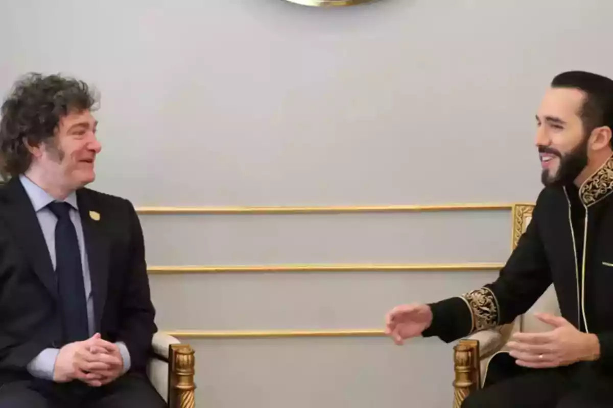 Dos hombres elegantemente vestidos conversan sentados en sillones dorados frente a una pared clara Dos hombres elegantemente vestidos conversan sentados en sillones dorados frente a una pared clara