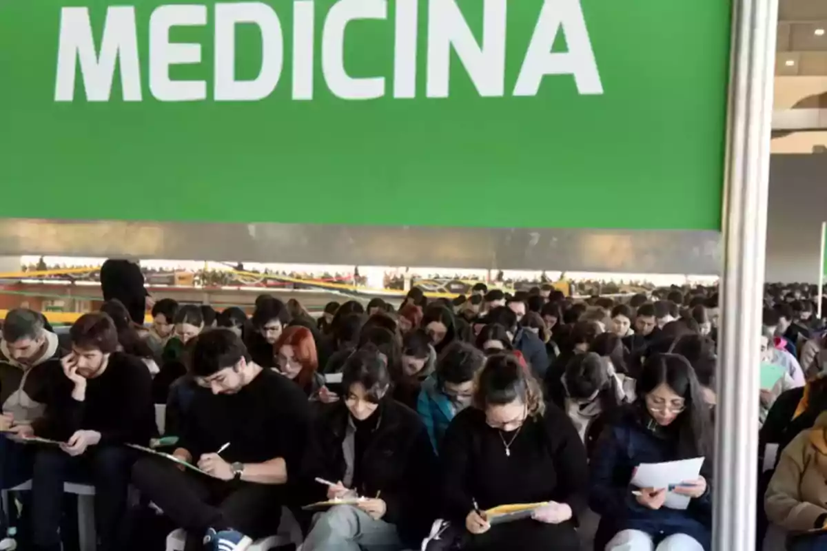 Un gran grupo de estudiantes sentados tomando un examen bajo un cartel verde que dice MEDICINA Un gran grupo de estudiantes sentados tomando un examen bajo un cartel verde que dice MEDICINA