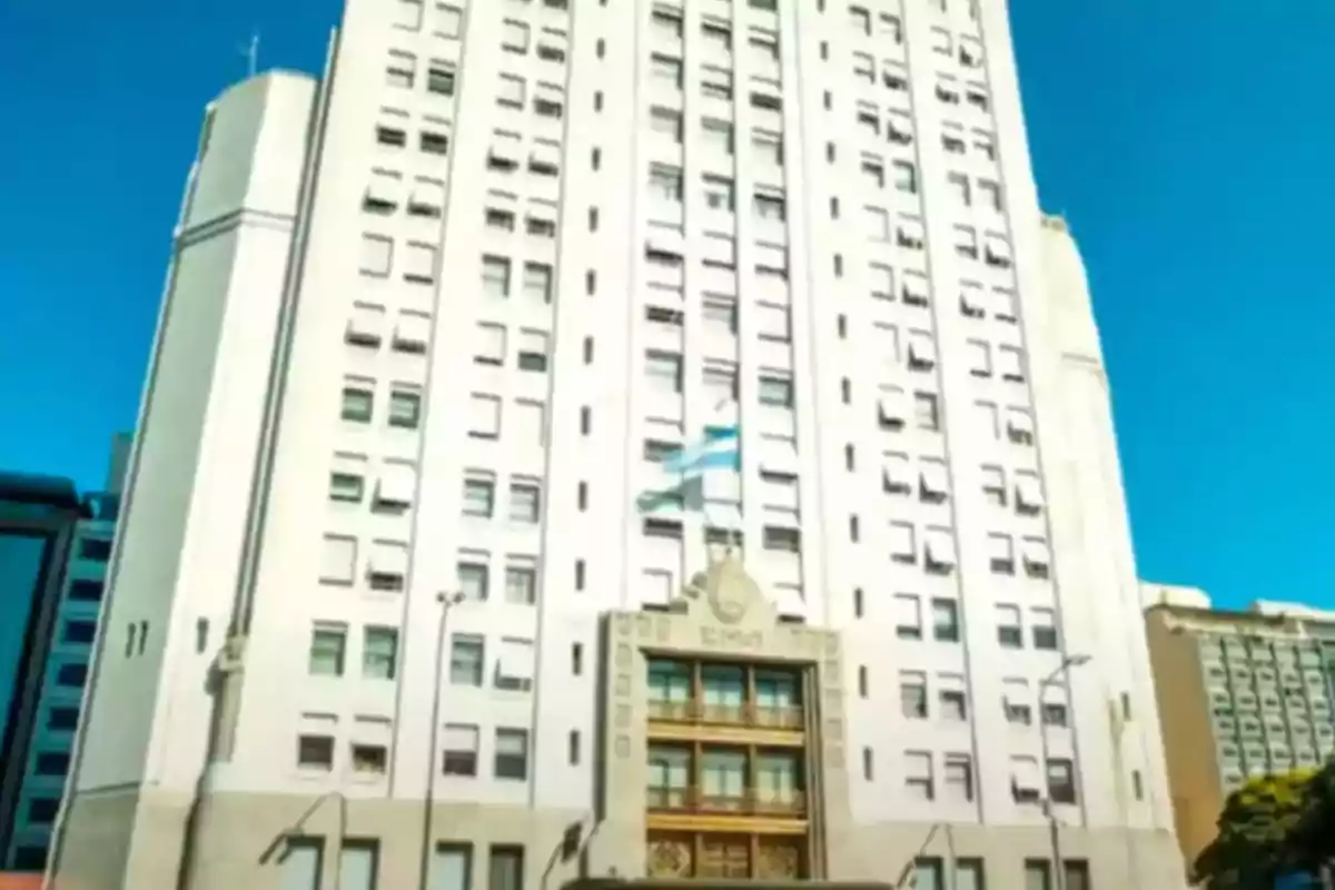 Edificio alto de color blanco con muchas ventanas y una bandera argentina ondeando en el centro bajo un cielo despejado Edificio alto de color blanco con muchas ventanas y una bandera argentina ondeando en el centro bajo un cielo despejado
