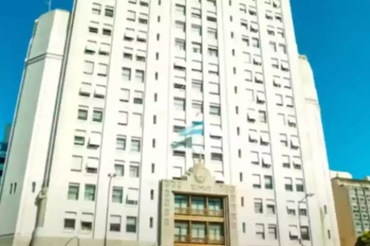 Edificio blanco de varios pisos con una bandera de Argentina ondeando en el centro y cielo azul de fondo Edificio blanco de varios pisos con una bandera de Argentina ondeando en el centro y cielo azul de fondo