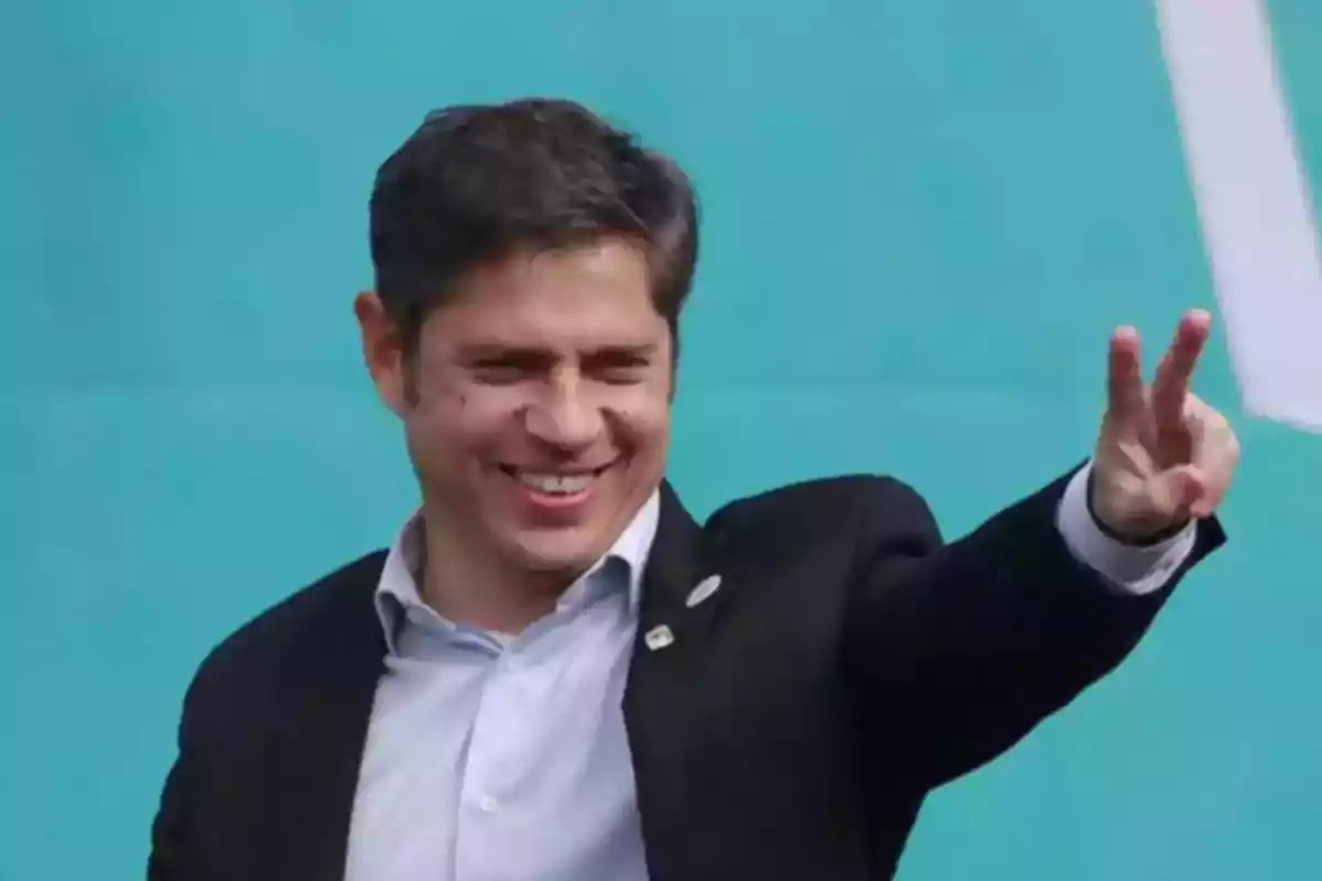 Hombre sonriente con traje haciendo el gesto de la victoria con la mano frente a un fondo celeste Hombre sonriente con traje haciendo el gesto de la victoria con la mano frente a un fondo celeste