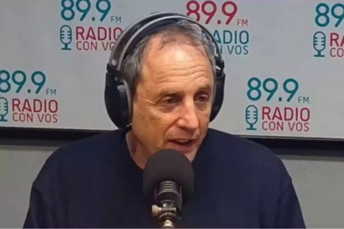 Older man wearing headphones and speaking in front of a microphone in a radio booth with the 89.9 FM Radio Con Vos logo in the background