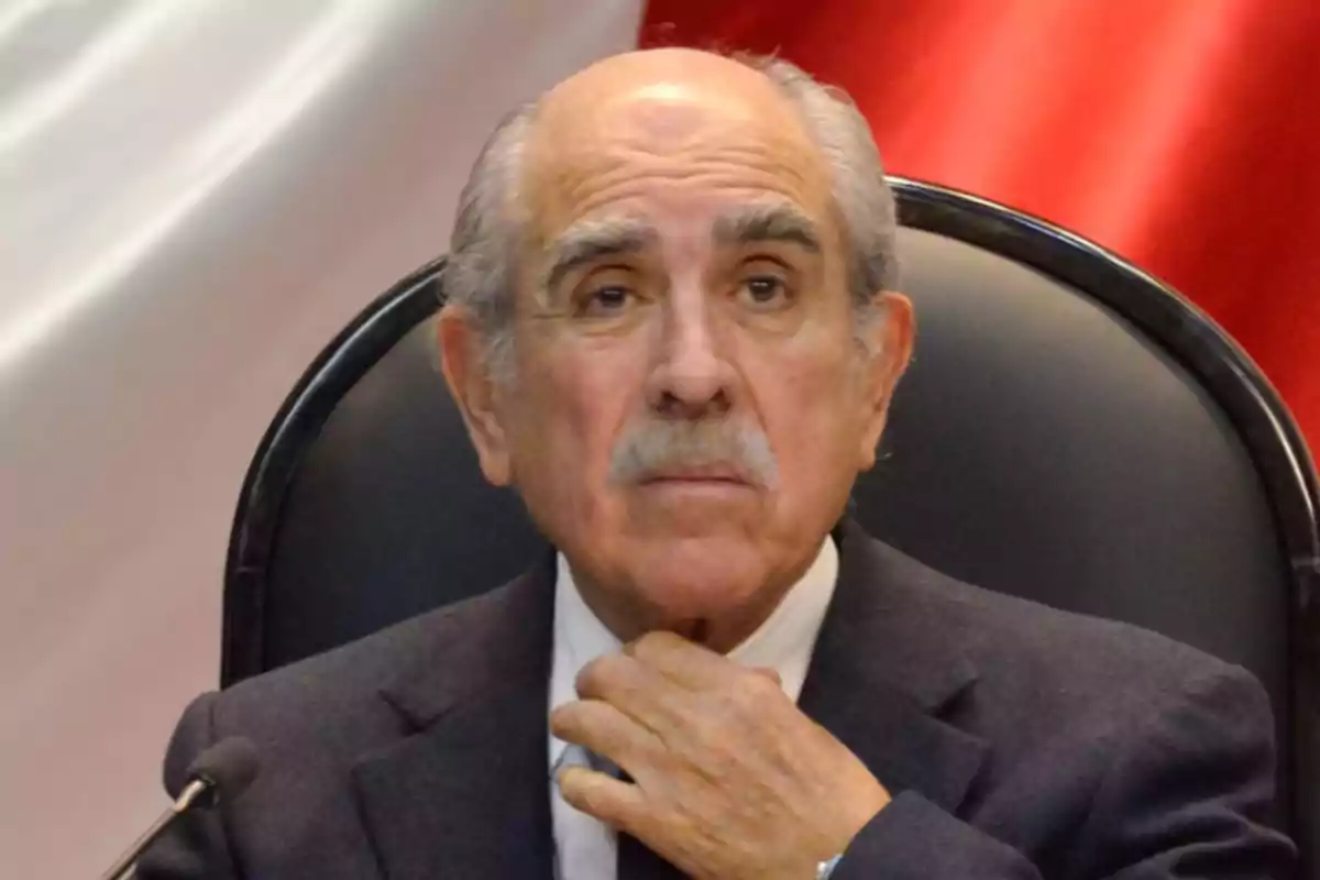 Hombre mayor de cabello canoso y bigote sentado en una silla negra con fondo de colores blanco y rojo