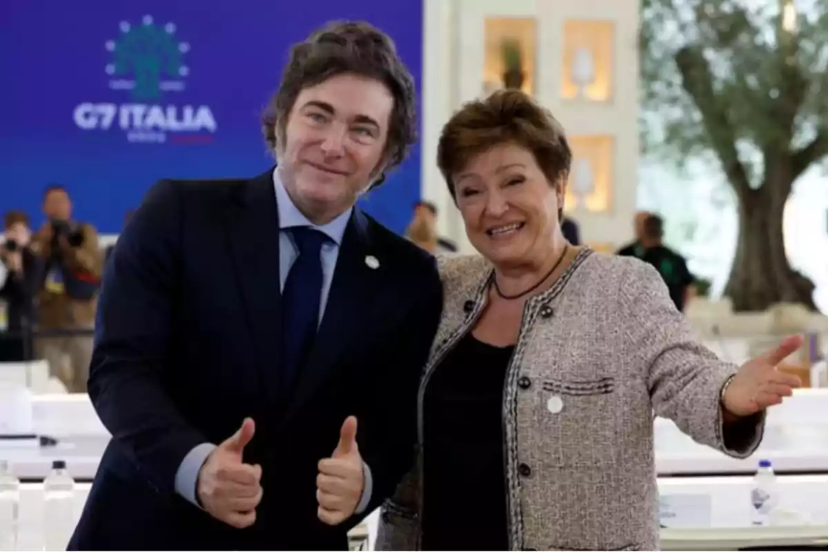 Dos personas sonríen y posan juntas en un evento formal, una de ellas hace el gesto de pulgares arriba y al fondo se observa un cartel con el texto G7 Italia.