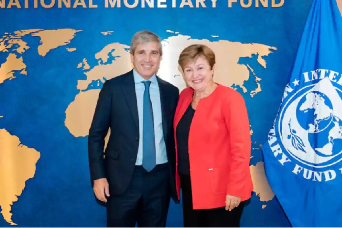Dos personas posan sonrientes frente a un fondo azul con un mapa dorado del mundo y una bandera del Fondo Monetario Internacional Dos personas posan sonrientes frente a un fondo azul con un mapa dorado del mundo y una bandera del Fondo Monetario Internacional