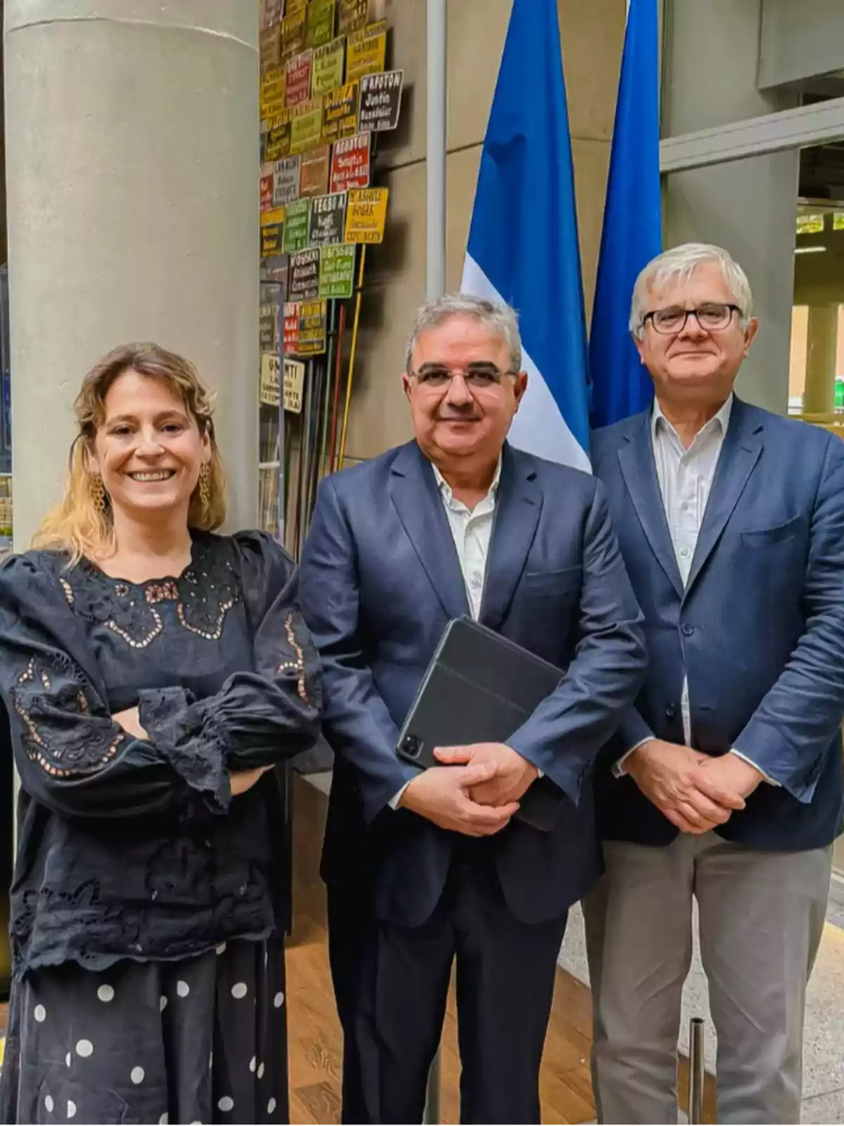 Tres personas posan sonrientes frente a dos banderas y un muro decorado con letreros de colores, todas vestidas de manera formal Tres personas posan sonrientes frente a dos banderas y un muro decorado con letreros de colores, todas vestidas de manera formal
