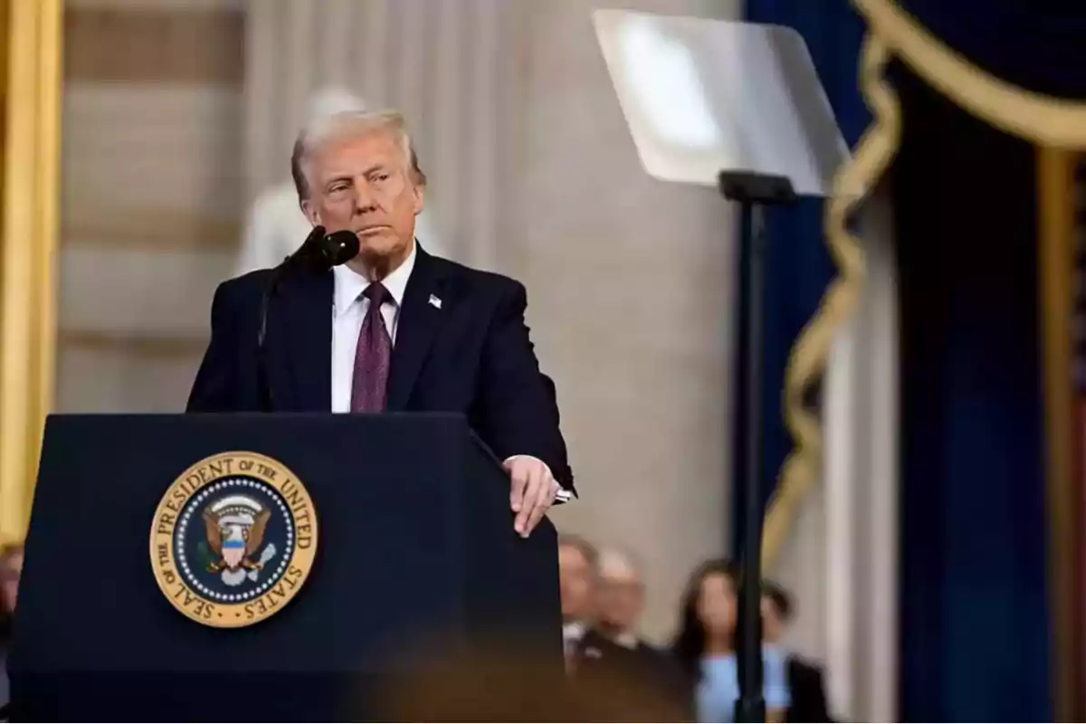 Hombre de cabello rubio con traje oscuro hablando en un podio con el escudo presidencial de Estados Unidos en un entorno formal Hombre de cabello rubio con traje oscuro hablando en un podio con el escudo presidencial de Estados Unidos en un entorno formal