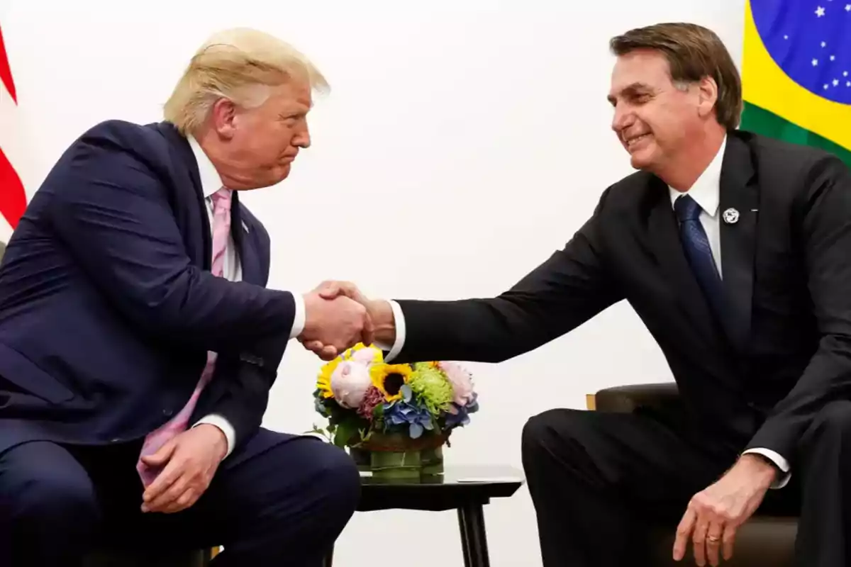 Dos hombres de traje se dan la mano sentados frente a una mesa con un arreglo floral, al fondo se ve una bandera de Brasil.