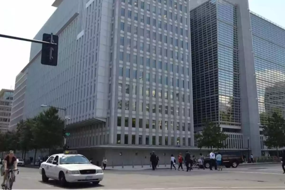 Edificio moderno de oficinas con fachada de vidrio y concreto en una esquina transitada donde personas cruzan la calle y un taxi blanco circula junto a un ciclista Edificio moderno de oficinas con fachada de vidrio y concreto en una esquina transitada donde personas cruzan la calle y un taxi blanco circula junto a un ciclista