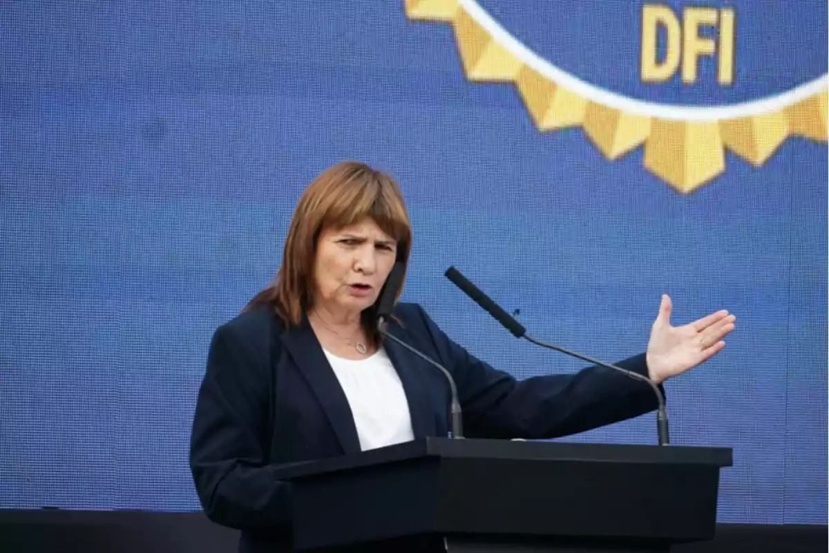Mujer de cabello castaño hablando en un podio con micrófono y fondo azul con letras amarillas