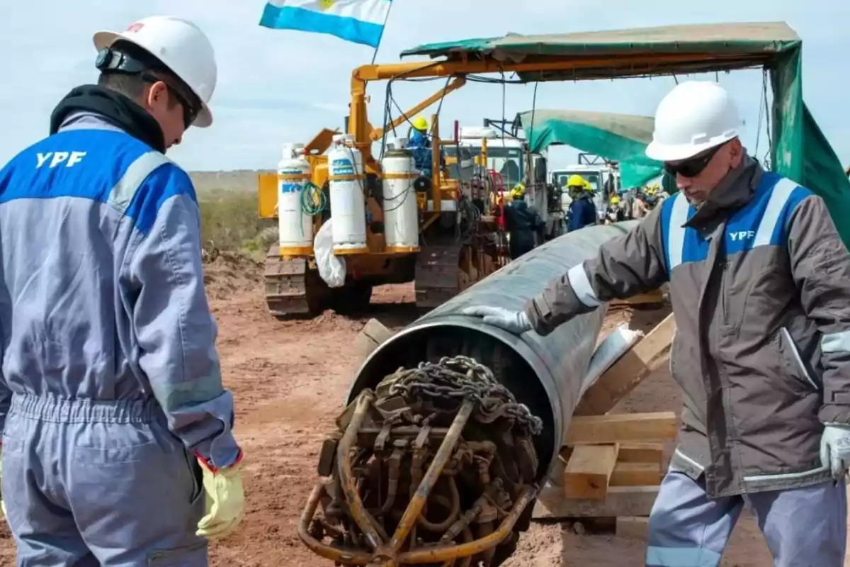 Workers wearing YPF uniforms and helmets supervise the installation of a gas pipeline at an outdoor construction site with heavy machinery and an Argentine flag in the background. Workers wearing YPF uniforms and helmets supervise the installation of a gas pipeline at an outdoor construction site with heavy machinery and an Argentine flag in the background.