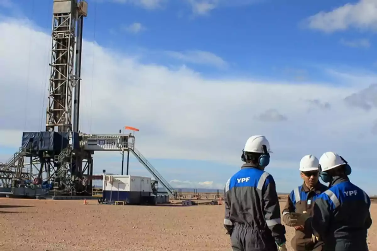 Tres trabajadores con cascos y uniformes de YPF conversan en un yacimiento petrolero al aire libre junto a una torre de perforación bajo un cielo parcialmente nublado